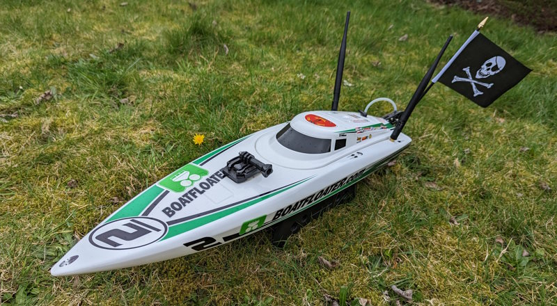 A model RC boat sat on a lawn. It has two large antennas and a pirate flag.