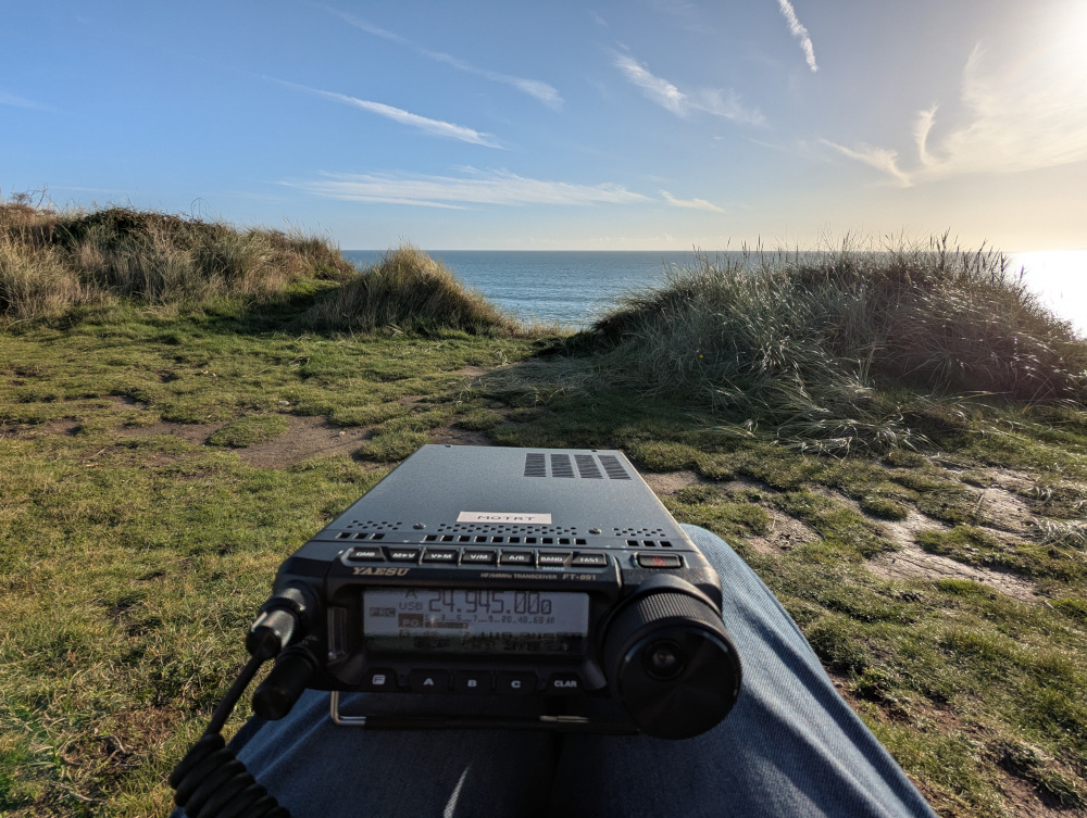 Radio in the foreground tuned to the 12m band, background of clifftop vegetation, sky and sea