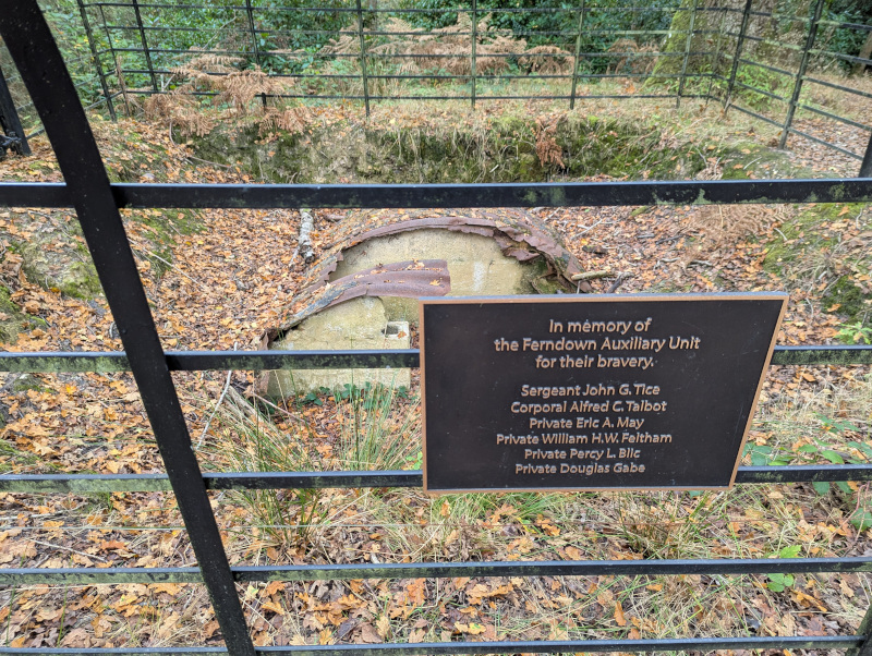 The concrete and steel ruins of a bunker. In the foreground, a fence bears a plaque with the names of the soldiers who served there.