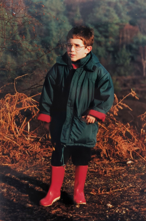 A roughly 6 year old me, wearing a big green coat and red boots amongst a brown autumnal background