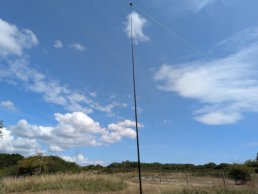 A vertical pole supporting a thin wire antenna. The background is dry scrub, trees and a wide blue sky.