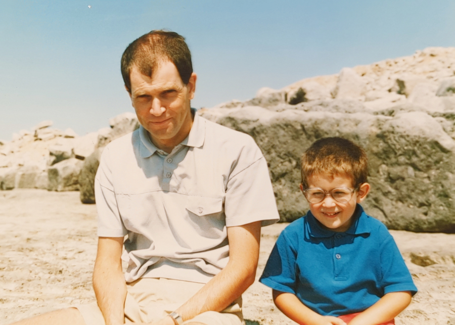 Three-year-old me in huge glasses, sat on a beach with my dad, with rocks behind and a blue sky