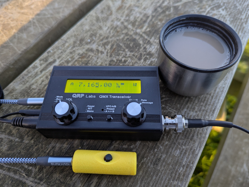 QMX transceiver, about the size of a pack of playing cards, on a picnic bench with a tiny mic and a cup of tea