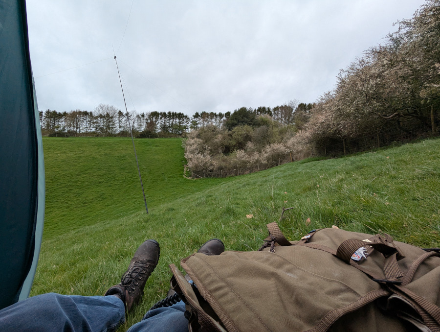 Photo taken looking out through the door of a tent. My feet are sticking out and a rucksack is lying on my leg. There is an antenna on a steep grassy slope in the background.